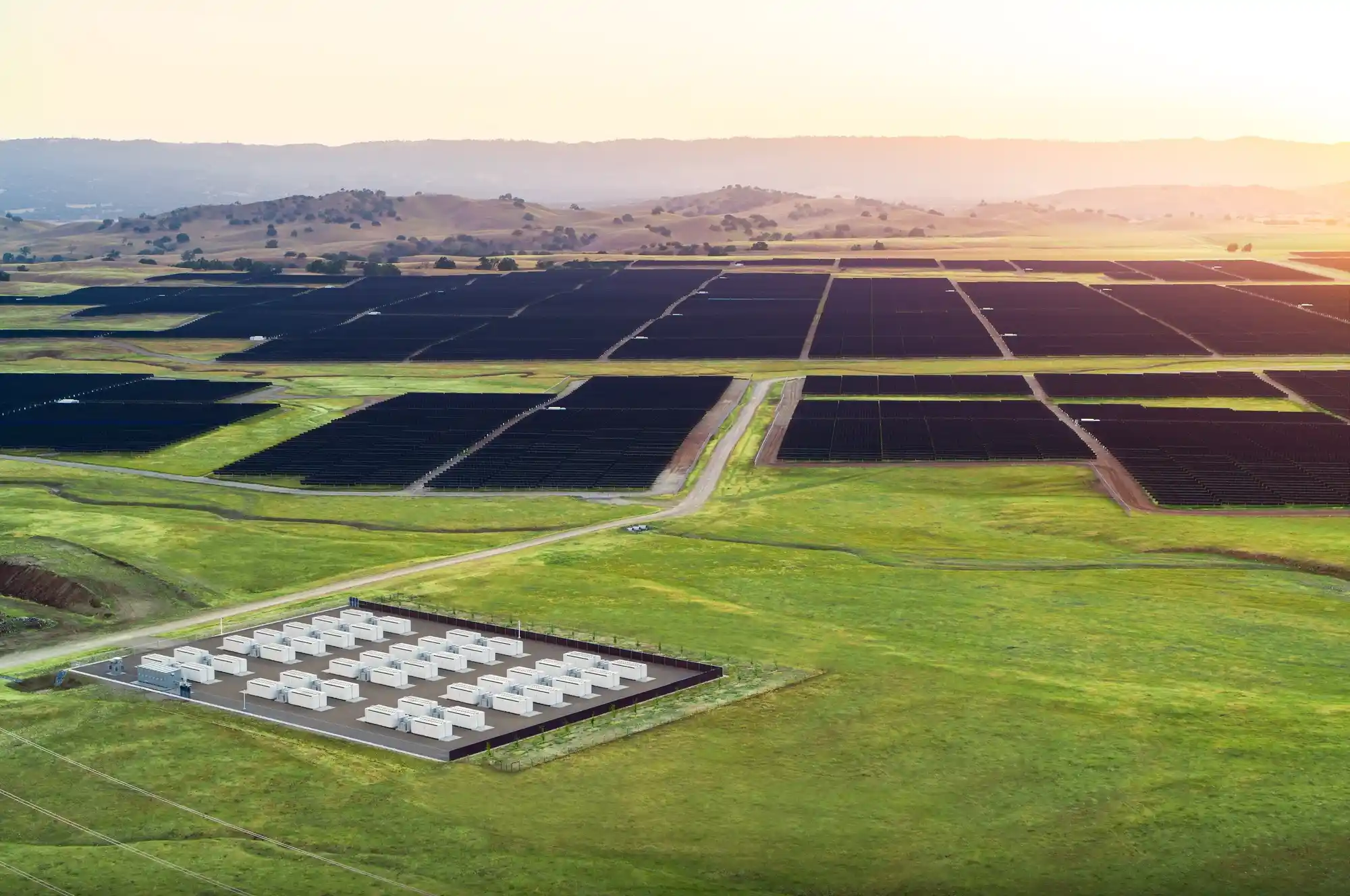 Aerial view of a commercial solar installation with large rectangular solar panels spread across fields. A small section of white structures is visible in the foreground. Rolling hills are seen in the background under a clear sky at sunset.