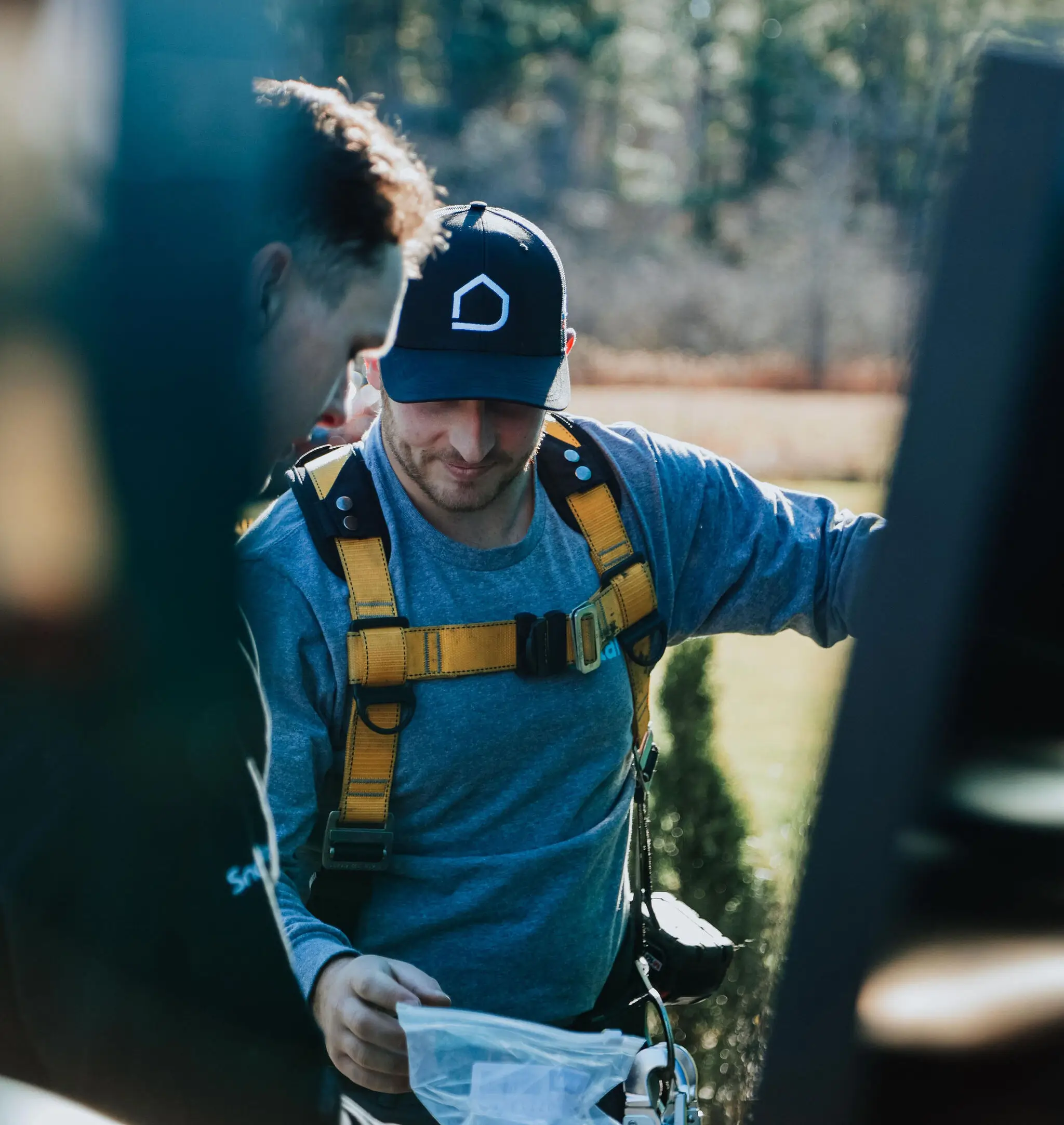Two men, one in a cap and yellow safety harness, review a document outdoors near solar panels on a sunny day, with blurred greenery in the background.