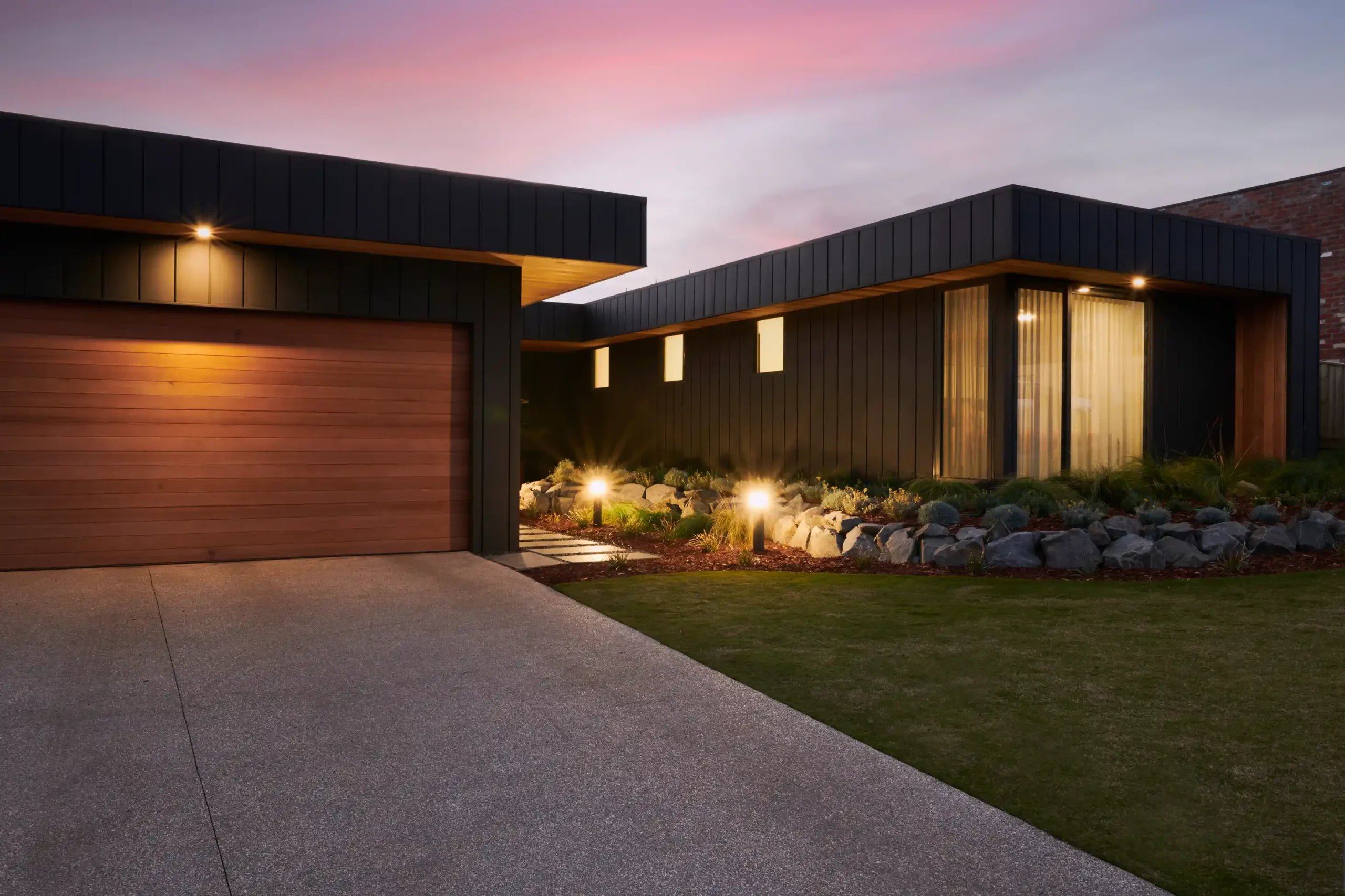A modern, single-story house with dark exterior walls and a wooden garage door is illuminated by outdoor lights powered by solar panels.