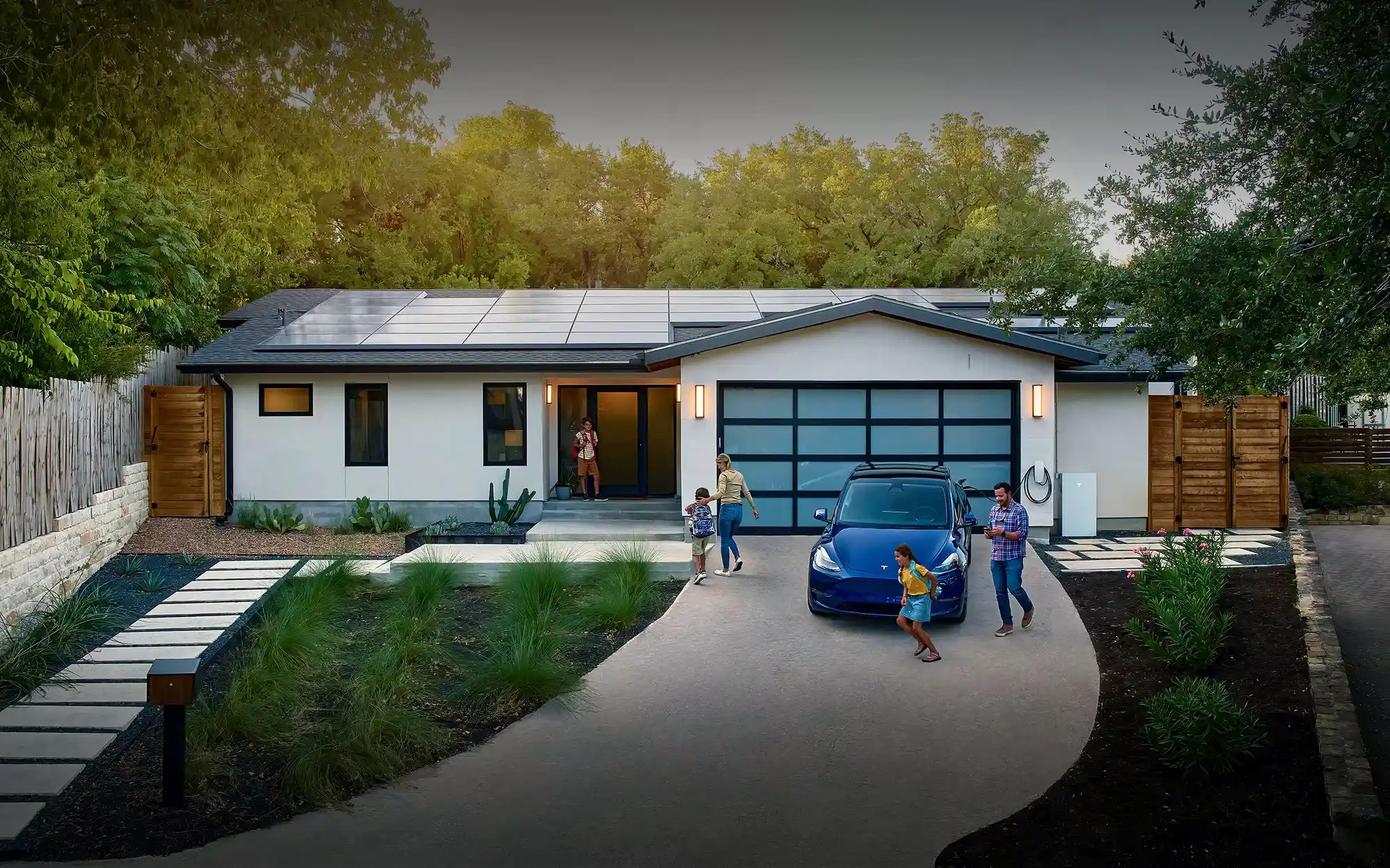 A modern, single-story house with solar panels on the roof and a glass garage door. A family of four stands near a parked blue car on the driveway.