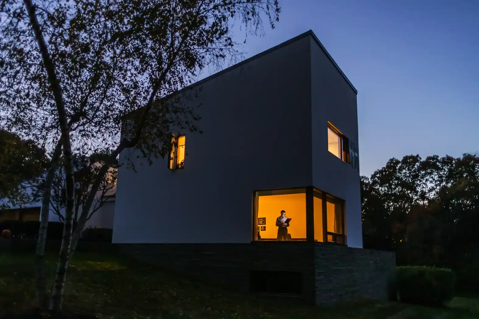 A modern white solar house at dusk with warm yellow lights glowing from the windows; a person stands inside visible through a large window.