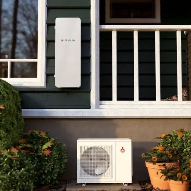 A Span electrical panel is mounted on a green house wall above a Mitsubishi outdoor HVAC unit, with bushes, a porch railing nearby, and solar potential for efficient energy management.
