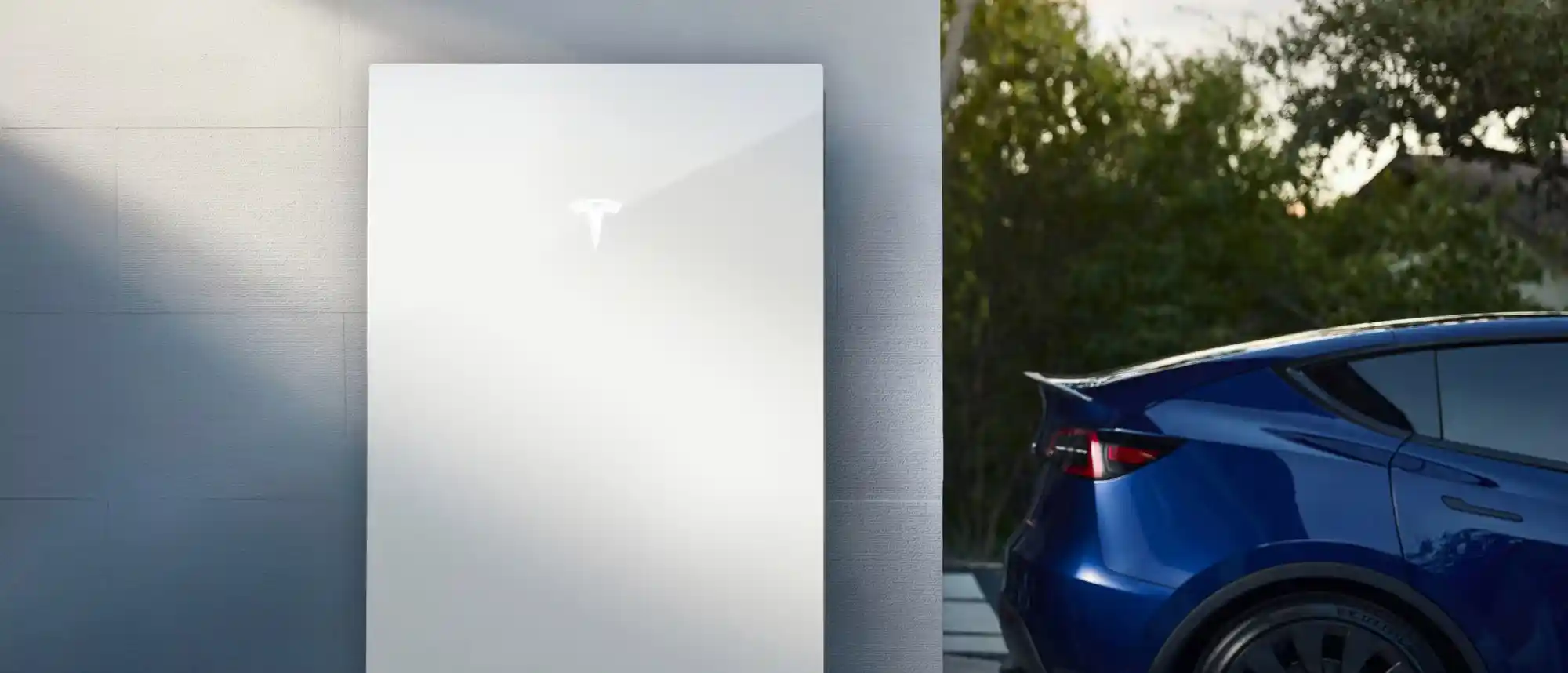 A white Tesla Powerwall battery is mounted on a light gray wall to the left. To the right, a blue car with a curved tail and spoiler is parked.