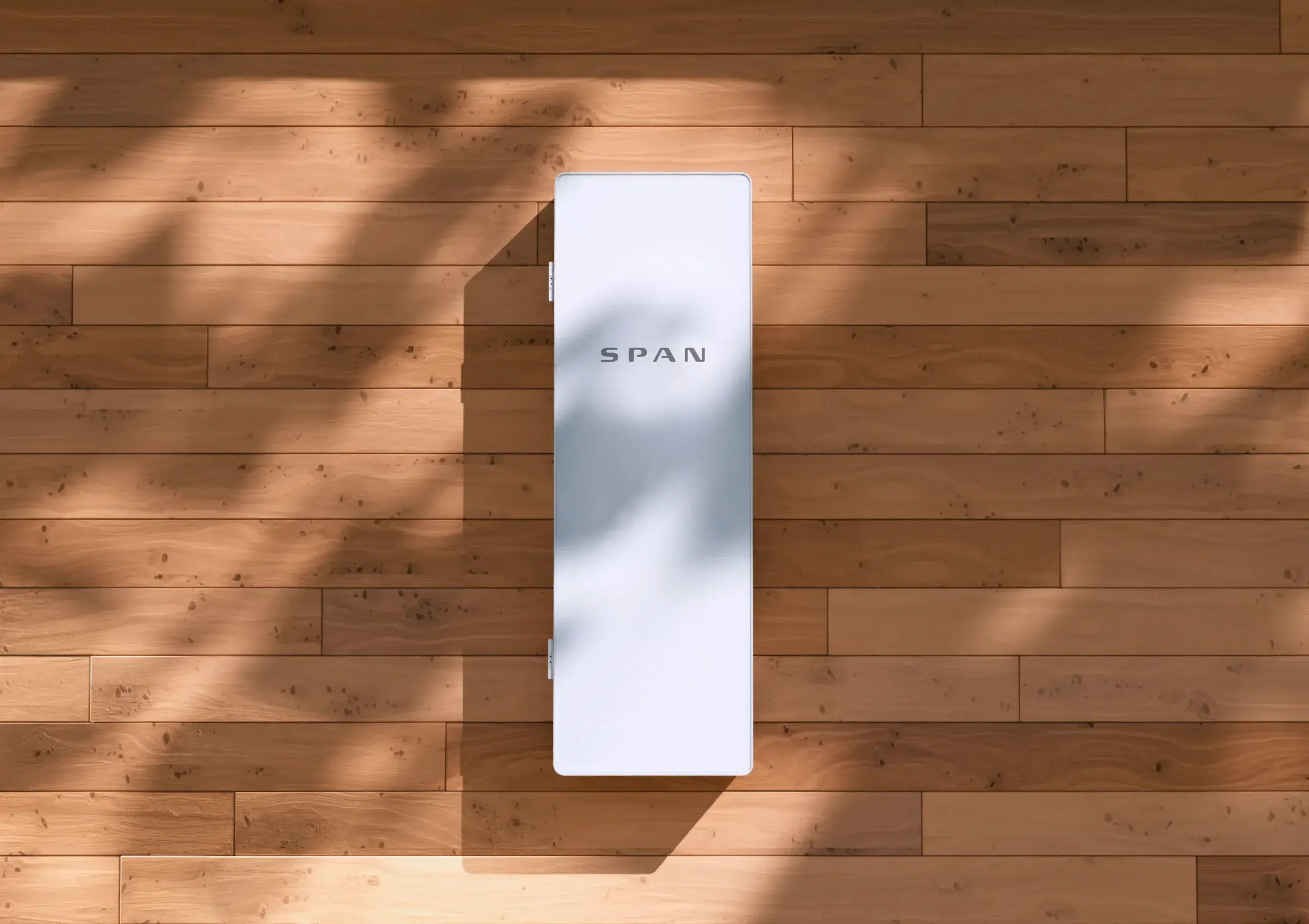 A white rectangular SPAN smart electrical panel mounted on a light wooden wall, with soft shadows of leaves cast across the surface.
