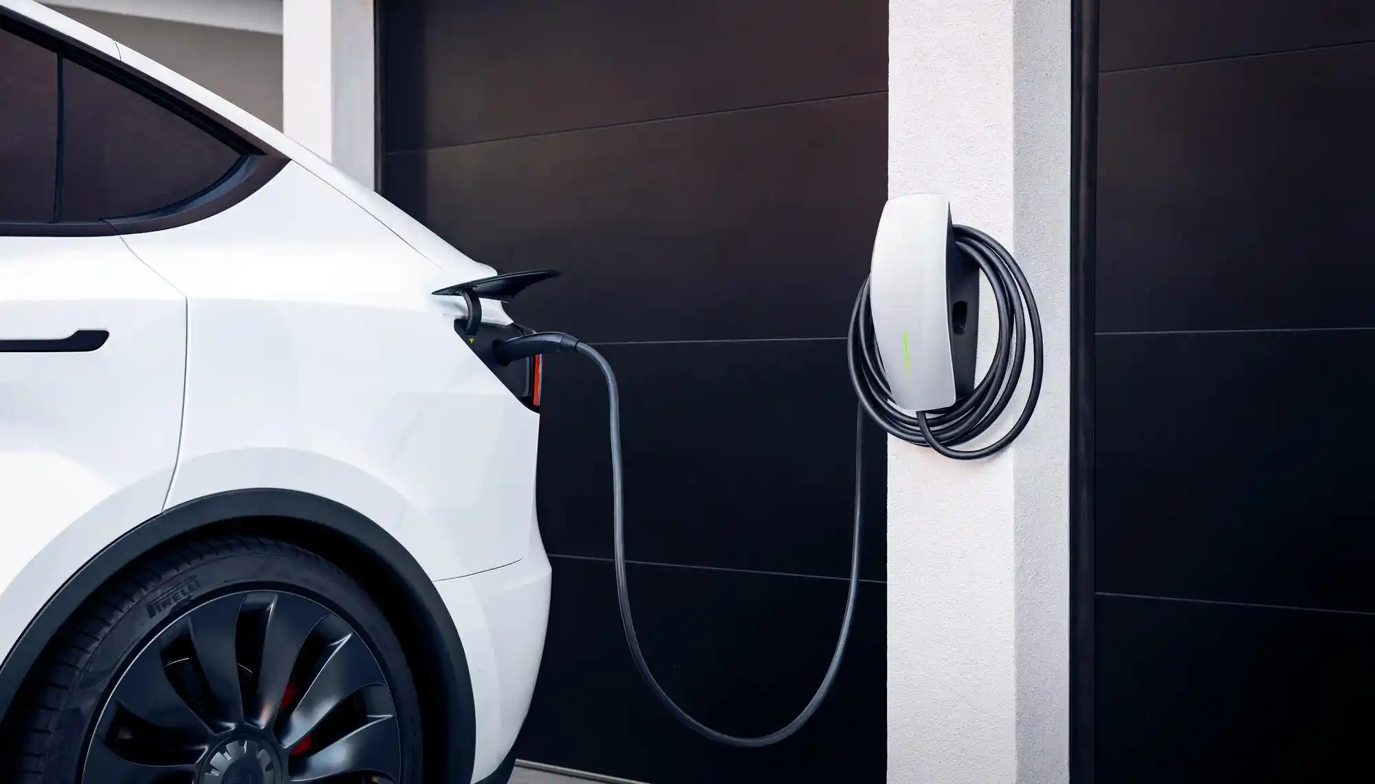 A white electric vehicle is parked and charging in a garage. The charging cable is plugged into the cars side port and connected to a wall-mounted charging station.