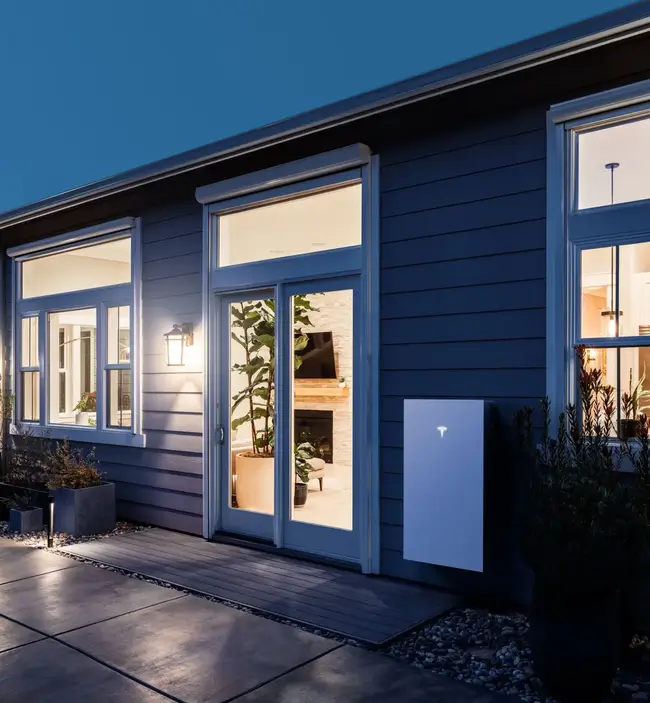 A modern house at dusk with large windows, warm indoor lighting, a potted plant inside, and a solar-powered Tesla Powerwall mounted outside on the wall near the door.