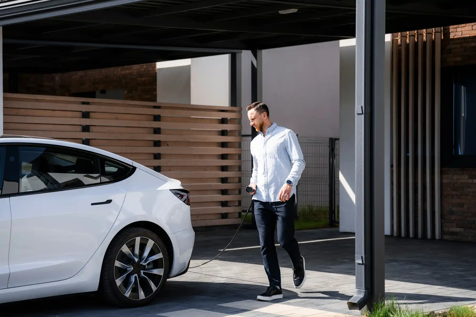 A man is walking towards a white electric car parked in a carport. He is holding a charging cable connected to the car.